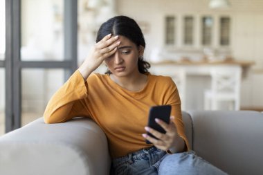 Evde koltukta oturmuş, akıllı telefon ekranına bakan ve kafasına dokunan genç Hintli kadın rahatsız edici bir mesaj okuyarak endişeli, kötü bir haber aldı, önemli bir telefon bekliyor.