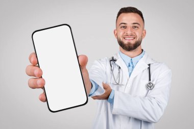 Genç Avrupalı doktor, boş ekranlı, gri arka planda ayarlanmış, sağlık hizmetleri için mükemmel bir cep telefonu sergiliyor.