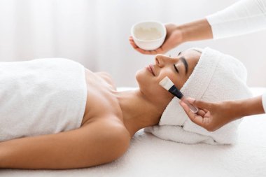 Yüz soyma maskesi, kaplıca güzellik tedavisi, cilt bakımı. Rahat genç Hintli kadın güzellik salonunda güzellik uzmanının yüz bakımını alıyor. Güzellik uzmanı kadın yüzüne beyaz maske takıyor.