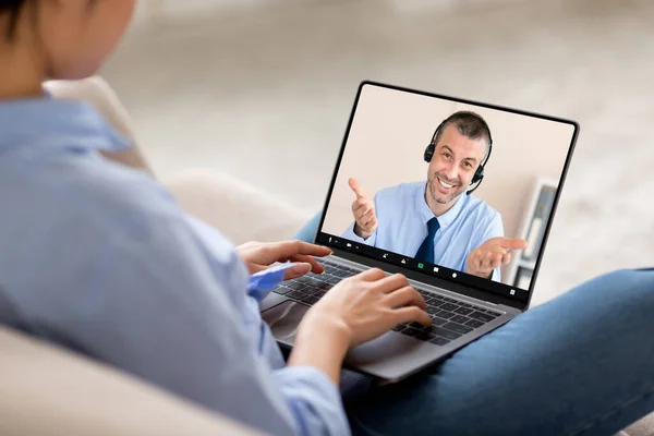 Over shoulder shot of young woman sitting on couch at home, using modern laptop, have video chat with mature man financial advisor, copy space