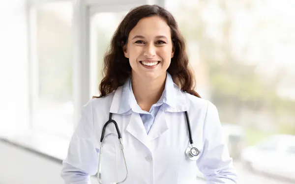 Cheerful millennial woman general physician standing by window at hospital or clinic, smiling at camera. Young brunette lady wearing white medical coat and carrying stethoscope smiling at camera