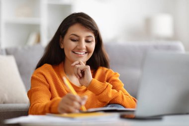 Yakın plan gülümseyen bin yıllık Hintli kız öğrenci öğretmeniyle online ders alıyor, oturma odasında oturuyor, dizüstü bilgisayar kullanıyor, not defterine notlar alıyor, ev içi, kopyalama alanı.