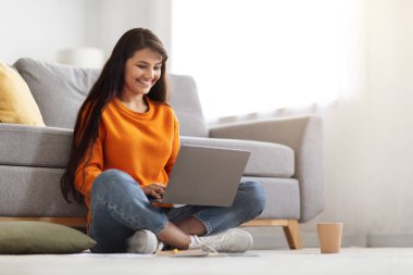Keyifli genç Hintli kadın, günlük kıyafetleriyle oturma odasında oturuyor, dizüstü bilgisayar kullanıyor, alışveriş yapıyor ya da online bankacılık yapıyor, İnternet 'te okuyor, web sörfü yapıyor, ev içini inceliyor, kopyalama alanı kullanıyor.