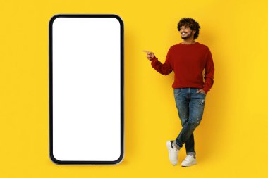 Güzel online teklif. Pozitif neşeli, yakışıklı Hintli genç adam beyaz ekranlı akıllı telefonu işaret ediyor, sarı stüdyo arka planında izole edilmiş, taklit edilmiş, kopyalama alanı