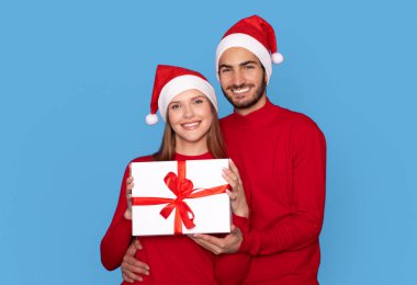 Noel Baba Şapkalı romantik çift paketlenmiş hediye kutusunu tutuyor ve kameraya gülümsüyor, mutlu Milenyum eşleri Noel tatillerinde karşılıyor, mavi stüdyonun arka planında tek başlarına duruyorlar,