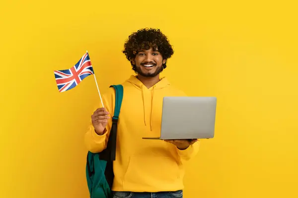 快乐的印度小伙子穿着黄色的帽衫背着背包 后面的学生拿着笔记本电脑和英国国旗 被黄色背景隔离 国外教育 学习英语概念 — 图库照片
