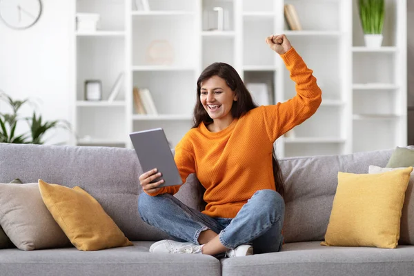 Excited young indian woman sitting on couch at home, using digital tablet, looking at gadget screen, raising hand up and screaming, got cashback, reading exciting news online, copy space