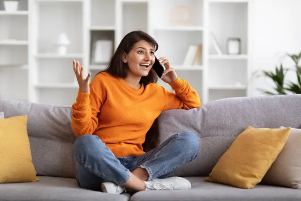 Emotional pretty young indian woman talking on phone at home. Happy hindu lady have conversation, using mobile phone, gesturing during funny call with boyfriend, looking at copy space. Communication