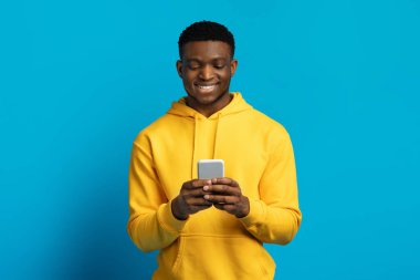 Halk ve Teknoloji Konsepti. Mavi stüdyo arka planında izole edilmiş akıllı telefon kullanan mutlu gülümseyen genç zenci portresi, fotokopi alanı. İnternette sohbet eden, sosyal medyada gezinen