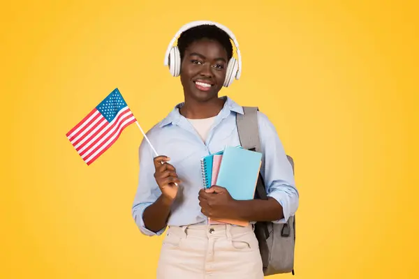 ワイヤレスヘッドフォンでハッピーな若いアフリカ系アメリカ人女性学生 アメリカの旗 イエロースタジオの背景に隔離された音楽を聴く 勉強と教育の交換 英語を学ぶ — ストック写真