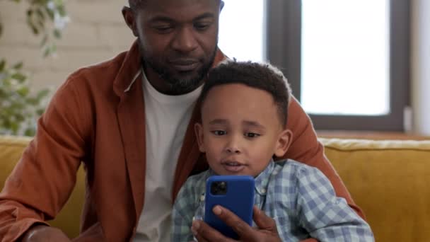 一緒に学ぶこと アフリカ系アメリカ人の父親と息子がスマートフォンでビデオを見たり 家で休んだり ショットを追跡したりする姿を閉じる ロイヤリティフリーのストック動画