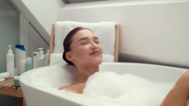 Yavaş çekim 4K video görüntüleri, lüks köpüklü bir banyoya dalmış mutlu ve keyifli bir kadını gösteriyor. Sahne huzur, rahatlama ve kendini pohpohlayan anların saf hazzını yayar..