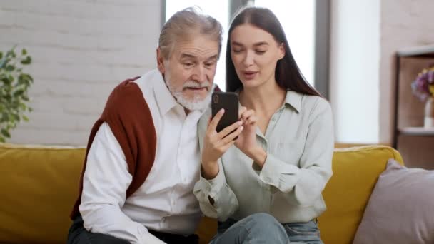 若い幸せな女性は 彼女の上級祖父にスマートフォンでモバイルアプリケーションを使用するように教え 自宅で一緒に時間を過ごし ビデオを見て笑う — ストック動画