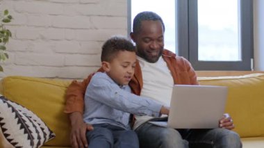 Modern uygun eğitim. Sevgi dolu babasıyla evde ders çalışan sevimli siyahi çocuk, dizüstü bilgisayardaki matematik ödevlerini çözüyor, online dersten zevk alıyor, çekim takibi, yavaş çekim, serbest alan.