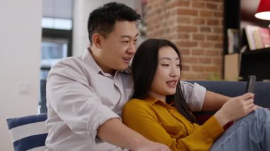 Gadget yaşam tarzı konsepti. Çinli evli çift akıllı telefon kullanıyor, hafta sonları dinleniyor, evdeki kanepede kaynaşıyorlar, birlikte vakit geçiriyorlar, çekim takip ediyorlar, yavaş çekim yapıyorlar.