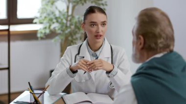 Tıp ve sağlık hizmetleri kavramı. Pozitif profesyonel kadın doktor randevu sırasında tanınmayan yaşlı bir adamla konuşuyor, ofisinde oturuyor, çekim yapıyor, yavaş çekim yapıyor.