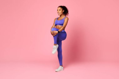 Antrenman ve sağlık. Gülümseyen Afro-Amerikan kadın spor kıyafetleri içinde kardiyo antrenmanına hazırlanıyor, bacak kaslarını esnetiyor pembe stüdyo arka planında dizlerini göğsüne dayıyor. Tam uzunluk