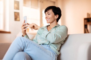 Orta yaşlı bir kadın akıllı telefonuna bakıyor, çevrimiçi sohbete dalmış, rahat bir şekilde evdeki rahat oturma odasında oturuyor, modern iletişim kavramını sergiliyor.