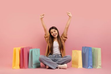 Pembe Stüdyo 'nun arka planında parlak alışveriş çantalarının yanında oturan neşeli genç kız, gülümseyen genç kız moda satışlarından zevk alıyor, ellerini kaldırıyor ve kameraya bakıyor, uzay boşluğunu kopyalıyor.