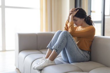 Depression Concept. Portrait Of Sad Young Indian Woman Sitting On Couch At Home, Lonely Eastern Female Feeling Upset And Desperate, Touching Head With Hands, Crying In Living Room, Copy Space clipart