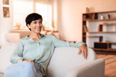 Esmer kadın gündelik oturma odasında kanepede oturuyor, evde dinlenirken mutluluk ve başarı yayıyor, hafta sonu keyfi ve konfor konseptinin resmi.