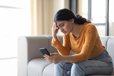 Haberler kötü. Evinde akıllı telefondan mesaj okuyan üzgün Hintli kadın endişeli, koltukta oturan ve dokunan kafaya dokunan, boşluğu kopyalayan cep telefonu ekranına bakan depresif bir kadın.