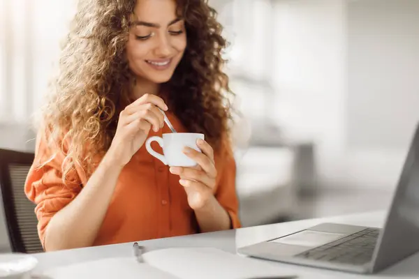 Curly-haired woman in orange blouse smiling and stirring cup of coffee, enjoying break while working on laptop in light, modern office setting, free space