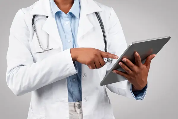 Millennial black woman doctor therapist in white coat, chatting at tablet, typing on gadget, isolated on gray background studio, cropped. Health care app, modern medicine