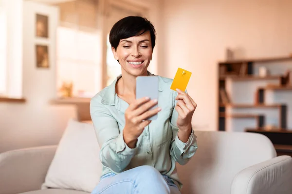 Cheerful middle aged lady holds smartphone and credit card, managing finances and shopping online on sofa in modern living room at home, browsing sales on cellphone and making secure payments