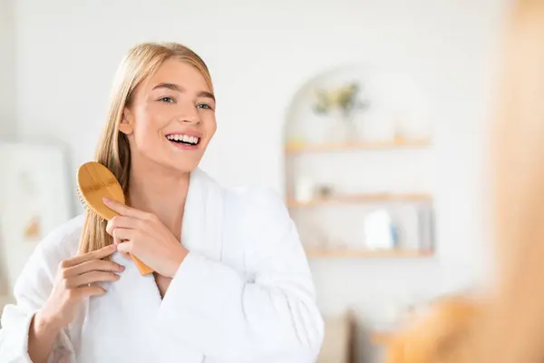 Young blonde lady in bathrobe brushing her long hair, standing in home bathroom interior, embodying daily wellness ritual and beauty routine, posing with wooden hairbrush