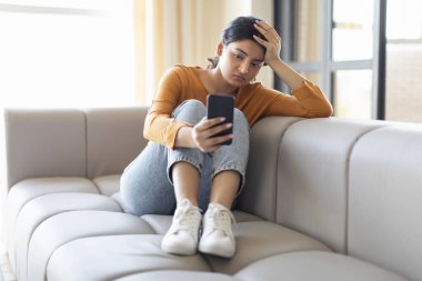 Smartphone ekranına bakan üzgün Hintli kadın, evinde koltukta oturan üzgün Doğu 'lu bayan, önemli bir telefon ya da mesaj bekliyor, mevsimsel depresyon geçiriyor, yalnız ve kalbi kırık.