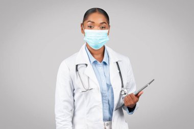 Beyaz önlüklü, koruyucu maske tutucu tabletli, gri arka plan stüdyosunda izole edilmiş gülümseyen siyahi doktor kadın terapist. Uzaktan sağlık uygulaması, danışma, muayene