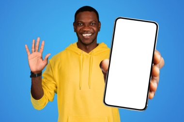 Genç Afrikalı Amerikalı bir adam elinde boş ekranlı büyük siyah bir cep telefonu tutuyor, merhaba diyor, uygulama ve teknoloji kavramları için ideal, mavi stüdyo arka planına karşı.