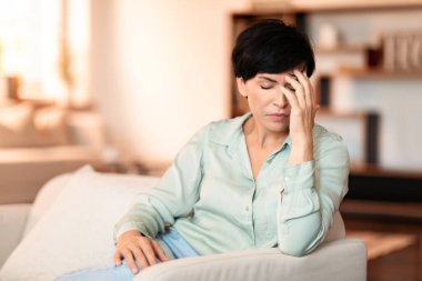 Orta yaşlı bir kadın başını koltukta acı içinde tutuyor, modern oturma odasında migren ağrısı ve stres tasviri, günlük hayatı etkileyen sağlık sorunları tasviri. Sağlık sorunları ve belirtileri