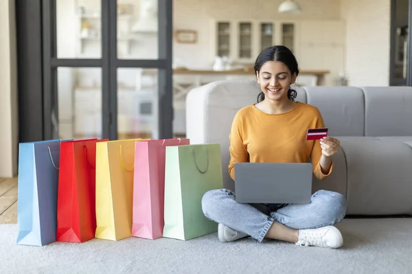 Online Shopping Concept. Beautiful Indian Woman Using Laptop And Credit Card At Home, Happy Eastern Female Sitting On Floor Next To Bright Shopper Bags, Enjoying Making Purchases In Internet