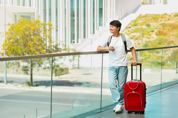Cheerful young asian man in casual outfit tourist with suitcase and phone in his hand standing outdoors next to airport or train station, waiting for taxi, looking at copy space