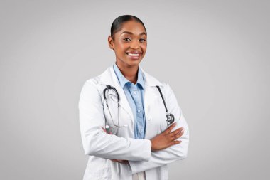 Profesyonel neşeli siyah doktor kameraya gülümsüyor, kolları bağlı, beyaz önlüklü ve steteskoplu, gri arka planda, stüdyoda. Sağlık, iş ve terapist yardımı