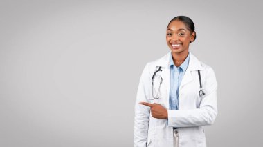 Gülümseyen profesyonel siyahi doktor uzayı işaret ediyor, sınav öneriyor, satış öneriyor, gri arka planda izole edilmiş, panorama. Dikkat dikkat, sağlık hizmeti tavsiyesi