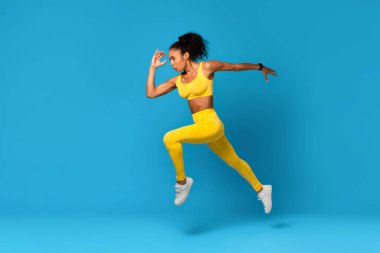 Motivasyonlu Afro-Amerikan Kadın Sarı Spor Giysiler Giyiyor Mavi Stüdyo Arkaplanı Üzerinde Orta Havada Atlama Egzersizi Yapıyor. Fitness Kadın Egzersiz Havada Sıçrıyor. Tam Uzunluk, Yan Görünüm