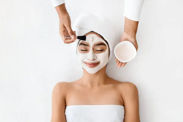 Top view of cosmetologist applying clay mask on face of young indian woman, beautiful eastern lady with towel on head lying on table, enjoying beauty treatments in spa salon, white background