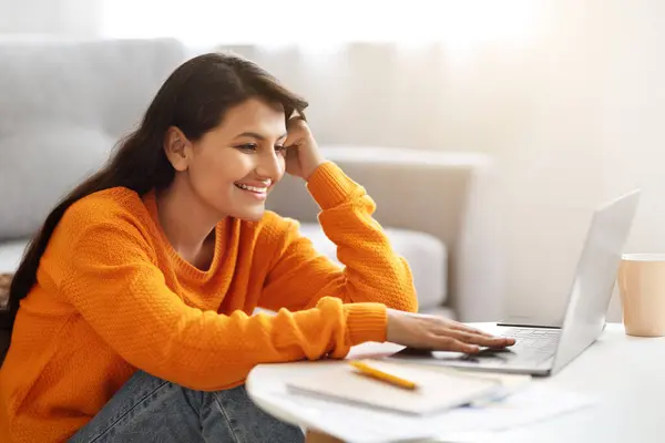 Closeup of positive beautiful long-haired young indian woman freelancer working from home, sitting on floor next to couch, typing on computer keyboard and smiling, copy space. Remote job