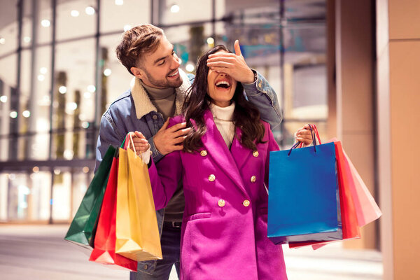 Веселый молодой человек удивительный женщина, закрыв глаза во время зимних покупок, пара за пределами магазина держат сумки, идеально подходит для отдыха коммерческие предложения, городской торговый центр на открытом воздухе в вечернее время