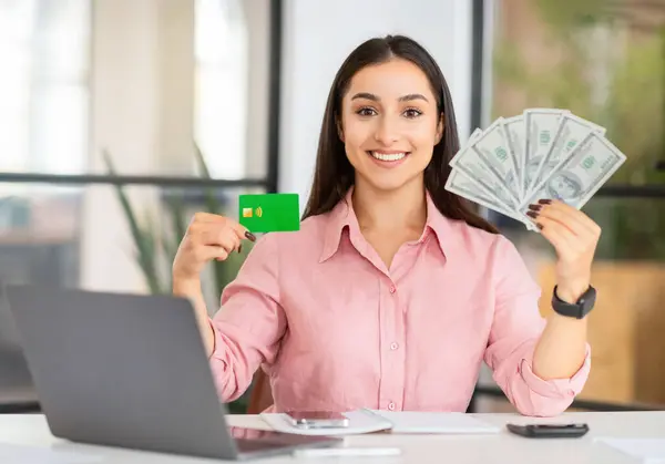 Happy young european woman manager at desk with laptop show credit card and many dollars cash in coworking office. Work, successful business recommendation, profit money, finance saving