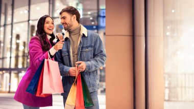 Neşeli alıcılar alışveriş merkezinin dışında gece alışveriş torbalarıyla poz veren, gülümseyen ve fotokopi alanına doğru hareket eden, tatil alışverişi için ideal bir çift. Panorama