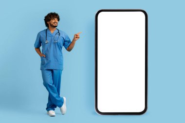 Mutlu, gülümseyen, tıbbi üniforma giyen yakışıklı Hintli doktor beyaz ekranlı büyük bir telefonu işaret ediyor, mavi stüdyo arka planı, model, teletıp konsepti.