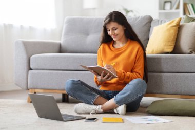Genç Hintli kadın çalışanlar online eğitim alıyorlar, ev içi. Milenyum hanımefendisi oturma odasında oturmuş not defterine notlar alıyor, dizüstü bilgisayarda ders dinliyor, boşluğu kopyalıyor.