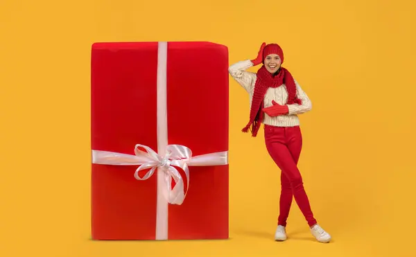 赤い編まれた帽子のハッピーな若い女性は 巨大な包まれたクリスマスの贈り物が笑顔で輝き 黄色の背景 完全な長さに対して撮影しました 冬のお祝いシーズンのセールス — ストック写真