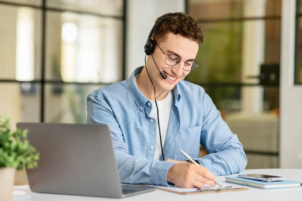 Smiling handsome young european man manager in casual with laptop at table with headphones taking notes in coworking office. Work, business, support remotely, call to client