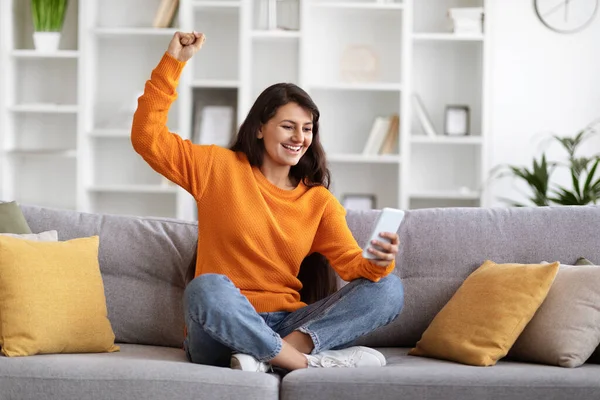 Excited young indian woman sitting on couch at home, using smartphone, looking at gadget screen, raising hand up and screaming, got cashback, reading exciting news online, copy space