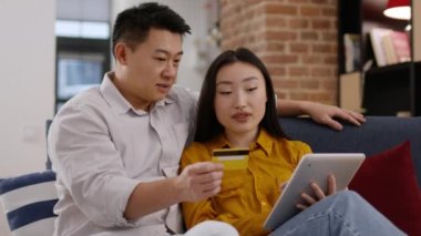 Çevrimiçi bankacılık kavramı. İnternette ödeme yapan mutlu Asyalı eşler, elinde kredi kartı olan bir adam ve dijital tablet uygulamasında veri dolduran bir kadın, izleme çekimi, yavaş çekim